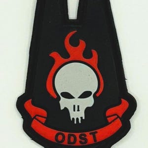 May include: Black patch featuring a white skull with red flames and a red banner that reads "ODST". The patch has a unique shape with two vertical extensions at the top.