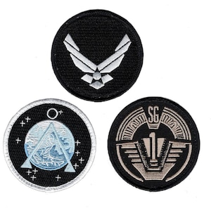 Puede incluir: Tres parches bordados circulares. El parche superior es negro con un logotipo plateado de la Fuerza Aérea. El parche inferior izquierdo es negro con un diseño triangular azul y blanco y pequeñas cruces. El parche inferior derecho es negro con texto SG-1 plateado.