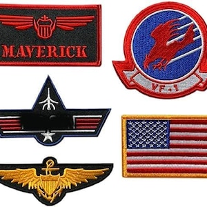 Puede incluir: Una colección de cinco parches bordados que incluye un parche rectangular negro con alas rojas y el texto MAVERICK, un parche circular azul con un águila roja y el texto VF-1, un parche triangular negro, un emblema de alas doradas y una bandera de EE. UU.