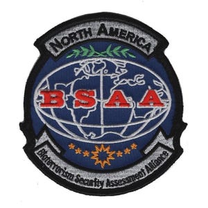 May include: A circular embroidered patch featuring a blue globe graphic with the red text BSAA across the center. The top banner reads NORTH AMERICA in black, while the bottom banner reads Bioterrorism Security Assessment Alliance in black.