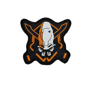 Puede incluir: Parche bordado con un cráneo blanco y naranja con espadas cruzadas. El diseño está sobre un fondo negro con detalles en naranja y azul. El parche tiene una forma irregular.