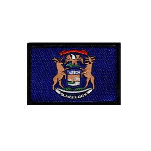May include: A rectangular embroidered patch featuring the Michigan state seal on a dark blue background. The design includes two tan deer standing on either side of a shield with the text TUEBOR, topped by an eagle and a red ribbon.