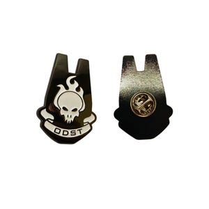 May include: Two black enamel pins. One pin features a white skull with flames and the text "ODST" on a banner. The other pin shows the back with a gold pin clasp. The pins are likely collectible items or accessories.