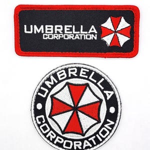 May include: A rectangular black embroidered patch with a red border featuring the white text UMBRELLA CORPORATION and a red and white umbrella logo, alongside a circular black patch with white text and a red and white umbrella emblem.