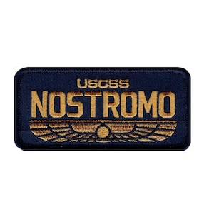 Op de afbeelding: Een rechthoekige geborduurde patch met een donkerblauwe achtergrond en een gouden rand. De tekst USCSS NOSTROMO is in gouddraad geborduurd boven een gestileerd gouden gevleugeld embleem met een centraal cirkelvormig detail aan de basis.
