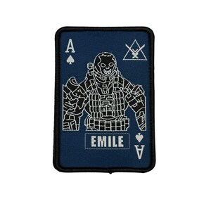 May include: A navy blue patch in the shape of a playing card, featuring a white illustration of a skull-faced soldier. The card is the Ace of Spades, with the word "EMILE" in white text.
