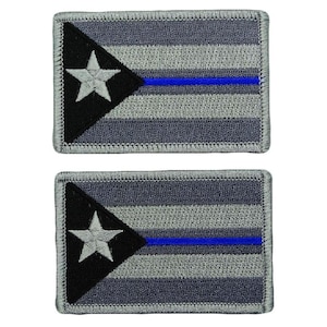 Puede incluir: Dos parches rectangulares bordados con un diseño de bandera en escala de grises. Cada parche tiene un triángulo negro a la izquierda con una estrella blanca, franjas horizontales grises y una fina línea azul en el centro.