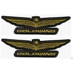 May include: Two identical embroidered patches featuring a stylized golden eagle with outstretched wings positioned above a black rectangular base. The word GOLDWING is stitched in gold capital letters across the center of the black base.