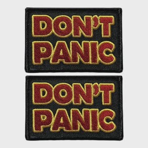 Könnte beinhalten: Zwei rechteckige schwarze Aufnäher mit den Worten "DON'T PANIC" in roter Farbe mit goldener Umrandung. Diese Aufnäher sind wahrscheinlich zum Aufnähen oder Aufbügeln auf Kleidung oder Taschen gedacht.