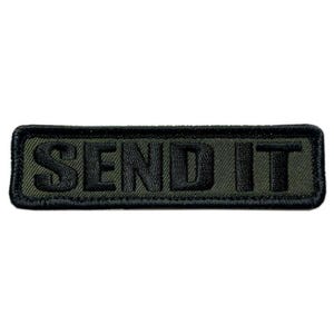 Puede incluir: Un parche rectangular bordado con un fondo verde oliva, un borde negro y el texto SEND IT cosido en letras mayúsculas negras y negritas en el centro. El parche tiene una apariencia de tela texturizada con costuras visibles.