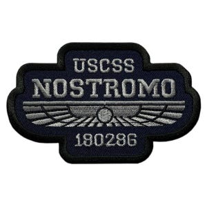 Può includere: Una toppa ricamata blu scuro con bordo nero. Il testo USCSS NOSTROMO e 180286 è ricamato in filo argentato. Un emblema centrale alato argentato con un cerchio al centro si trova tra le righe di testo, caratterizzato da cuciture dettagliate.