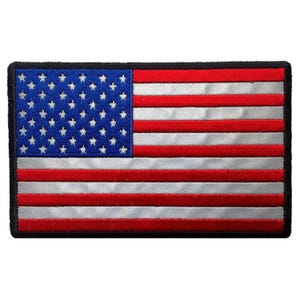 Puede incluir: Un parche rectangular bordado con la bandera de los Estados Unidos. El diseño incluye un cantón azul con estrellas blancas a la izquierda y franjas horizontales alternas de color rojo y blanco plateado a la derecha, con un borde negro.