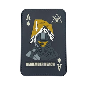 May include: A rectangular patch featuring a stylized design of a soldier's helmet and the text "REMEMBER REACH." The patch is in the shape of an Ace of Spades playing card, with a mountain and sword graphic.