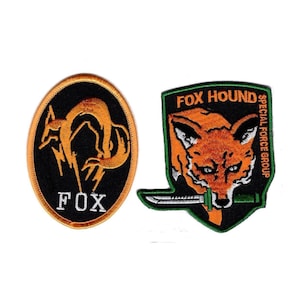 May include: Two embroidered patches. The left is an oval with a black background, an orange fox silhouette, and the text FOX. The right is a shield-shaped patch with a black background, an orange fox head, a green knife, and FOX HOUND SPECIAL FORCE GROUP.