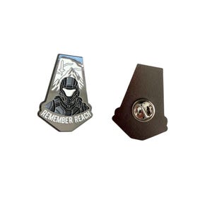 May include: A silver-colored enamel pin with a design featuring a helmeted figure, mountains, and the text "REMEMBER REACH." The pin has a black backing with a pin clasp.