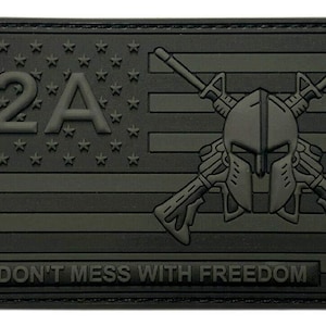Puede incluir: Un parche táctico rectangular negro con un diseño en relieve de la bandera estadounidense con el texto 2A en el cantón. Un casco espartano está centrado sobre dos rifles cruzados en las franjas, con el texto DON'T MESS WITH FREEDOM en la parte inferior.