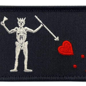 Op de afbeelding: Een rechthoekige zwarte geborduurde patch met een wit skeletfiguur dat een speer in de ene hand en een klein voorwerp in de andere hand houdt. Rechts van het skelet staat een rood hart, vergezeld van drie kleine rode stippen.