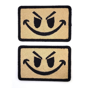 May include: Two rectangular embroidered patches featuring a tan background with a black stitched border. Each patch displays a black embroidered graphic of a mischievous face with two angled, sharp-edged eyes and a wide, curved, upward-turning grin.