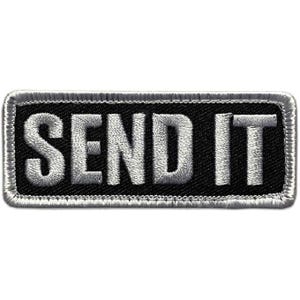 May include: A rectangular embroidered patch with a black background and a silver border. The text SEND IT is stitched in silver thread in a bold, sans-serif font across the center of the patch. The edges are finished with a silver merrowed border.