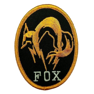 Puede incluir: Un parche bordado de forma ovalada con fondo negro y un gráfico de zorro estilizado en color dorado. El borde está cosido con hilo dorado y la palabra FOX está bordada en letras mayúsculas de color gris claro en la parte inferior.