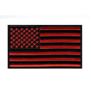May include: A rectangular embroidered patch featuring a stylized American flag design. The canton displays red stars on a black background, while the field consists of alternating horizontal red and black stripes. The edges are finished with black stitching.