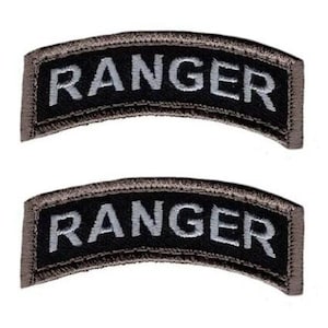 Pode incluir: Dois emblemas bordados curvos idênticos com fundo preto e a palavra RANGER bordada em letras maiúsculas cinza-claras. Cada emblema possui uma borda cinza-escura e um formato arqueado distinto para fixação em uniformes.