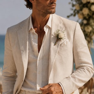 May include: A beige linen blazer and matching trousers worn with a white button-down shirt. The blazer features a notched lapel, a pocket square, and a small floral boutonniere with white flowers and greenery pinned to the left chest area.