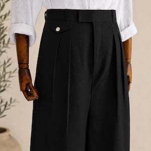 May include: Black high-waisted trousers with pleats and a wide waistband. The trousers have a buttoned pocket detail and are paired with a white button-down shirt. The pants are displayed on a wooden mannequin.