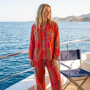 May include: A striking pajama set in red and orange, with a tiger stripe design. The long-sleeved top and trousers have a classic button-down style. The model is standing on a wooden deck with the sea in the background.