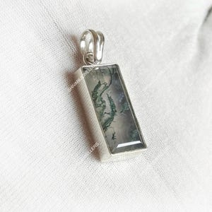 May include: A rectangular silver pendant with a moss agate stone. The stone features green and white patterns. The pendant has a loop at the top for a chain. The text "SHIVANI JEWELLERS" is visible on the side.