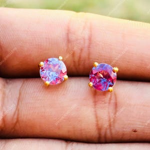 May include: A pair of gold-colored stud earrings, each featuring a round, faceted gemstone with a blend of pink and purple hues. The earrings are set with small gold prongs.