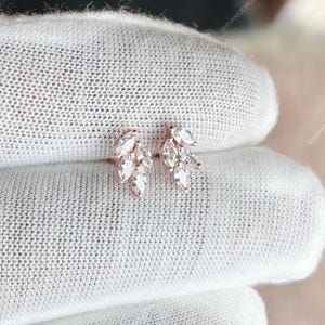Pode incluir: Brincos de tachas em ouro rosa. Cada brinco apresenta um grupo de pedras de zircónia cúbica transparente com corte marquesa, dispostas em forma de folha. Os brincos são apresentados sobre um fundo branco.