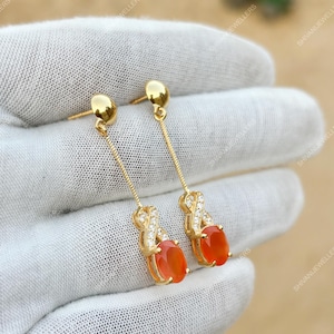 Könnte beinhalten: Ein Paar goldfarbene Ohrhänger. Jeder Ohrring hat einen Stecker, eine filigrane Kette, ein dekoratives Element mit kleinen, klaren Steinen und einen ovalen, orangeroten Edelstein. Die Ohrringe werden vor weißem Hintergrund gezeigt.