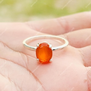 May include: A silver ring with an oval, orange-red gemstone. The gemstone is set in a polished silver band. The ring's design is simple, emphasizing the gemstone's colour. The ring is shown against a light background, enhancing its appearance.