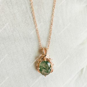 May include: Rose gold necklace with a pendant featuring a green and white oval gemstone. The pendant is encased in a decorative rose gold setting. The chain is delicate and the necklace is displayed on a white surface.