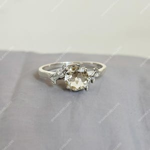 Peut inclure: Une bague en argent avec une grosse pierre précieuse claire. La bague présente un motif floral sur le bandeau, avec des feuilles et des détails délicats. La pierre précieuse est de forme ovale et sertie dans un sertissage à griffes. La bague est présentée sur une surface grise.