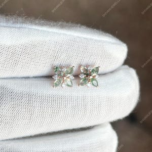 Puede incluir: Un par de pendientes de botón con un diseño floral. Cada pendiente tiene cuatro piedras verdes en forma de pétalo y pequeñas piedras redondas y transparentes. Los pendientes están engarzados en un metal de color oro rosa. Los pendientes se muestran sobre una superficie blanca.