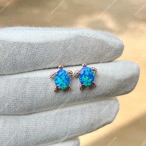 Könnte beinhalten: Ein Paar roségoldene Schildkröten-Ohrstecker mit leuchtend blauen Opalsteinen. Die Ohrringe sind auf einer weißen Oberfläche platziert, was das detaillierte Design und die Farbe der Edelsteine hervorhebt. Ein einzigartiges Schmuckstück.