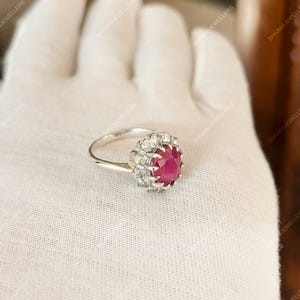 May include: A silver ring featuring a large, oval-cut ruby center stone surrounded by a halo of smaller, clear gemstones. The ring is set against a white background, showcasing the jewelry's details.