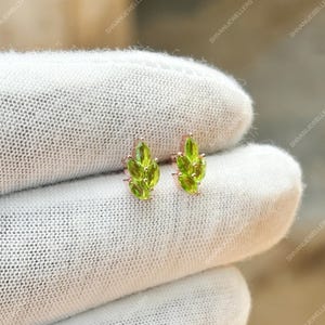 May include: A pair of stud earrings featuring a leaf design. Each earring has a cluster of bright green gemstones set in a rose gold metal. The earrings are displayed on a white fabric background.