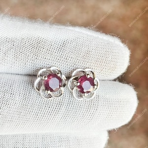 Pode incluir: Um par de brincos de botão prateados. Cada brinco apresenta uma pedra preciosa redonda, vermelha escura, engastada numa moldura em forma de flor. Apresentados sobre uma superfície branca e texturizada.