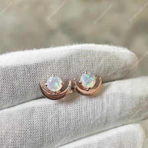 May include: A pair of rose gold earrings featuring a crescent moon design. Each earring has a round, iridescent opal stone set in the center, with a small accent stone. The earrings are displayed on a white surface.