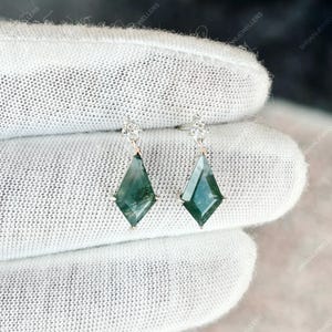 May include: A pair of silver earrings with a diamond-shaped, green gemstone pendant. The earrings are held against a white background. The gemstones have a unique, moss-like pattern. The earrings are a delicate piece of jewelry.