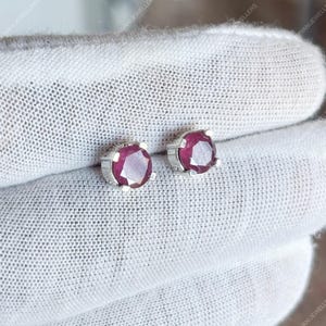 Może przedstawiać: Para srebrnych kolczyków sztyftów, każdy z okrągłym, fasetowanym, głęboko czerwonym kamieniem szlachetnym. Kolczyki osadzone są w prostej, czteroząbkowej oprawie, podkreślającej żywy kolor kamieni. Kolczyki są prezentowane na białej powierzchni.