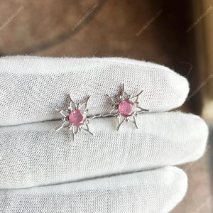 Puede incluir: Un par de pendientes de plata con cierre de tuerca. Cada pendiente presenta una piedra preciosa rosa en el centro, rodeada por un diseño de estrella. Los pendientes se muestran sobre una superficie blanca.