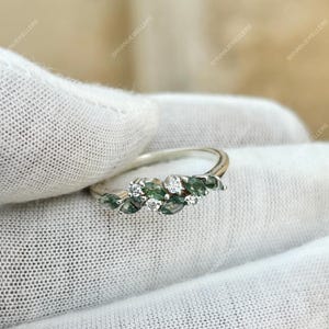 Puede incluir: Un anillo de plata con un grupo de piedras preciosas verdes y blancas. El anillo presenta piedras verdes con corte marquesa y piedras blancas redondas, engastadas en una banda delicada. El anillo se muestra sobre una superficie blanca.