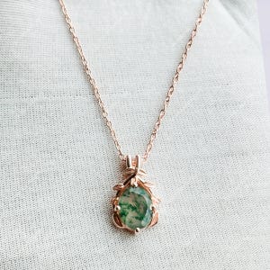 May include: Rose gold necklace with a pendant featuring a green and white stone. The pendant is surrounded by a decorative rose gold frame with leaf accents. The chain is delicate and the necklace is displayed on a white surface.