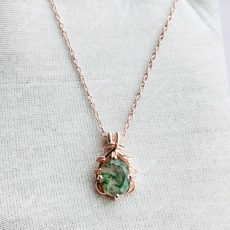 May include: Rose gold necklace with a pendant featuring a green and white stone. The pendant is surrounded by a decorative rose gold frame with leaf accents. The chain is delicate and the necklace is displayed on a white surface.