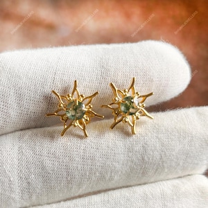 May include: A pair of gold-colored stud earrings. Each earring features a central, round, light green stone surrounded by a starburst design. The earrings are displayed on a white surface.