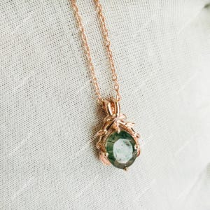 May include: Rose gold necklace featuring a round, green gemstone pendant. The pendant is set in a decorative frame with leaf-like accents. The chain is delicate and complements the pendant's design. The jewelry is displayed on a textured, off-white surface.